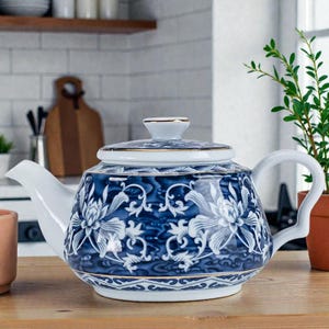 May include: A blue and white porcelain teapot with a floral design. The teapot has a white handle, spout, and lid. Gold trim accents the lid and base. The teapot is sitting on a wooden surface.