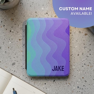Può includere: Una custodia personalizzata per e-reader con un motivo a onde nei toni del blu, verde e viola. Il nome "JAKE" è stampato in lettere scure in basso a destra. Il testo "CUSTOM NAME AVAILABLE!" è in un cerchio viola.