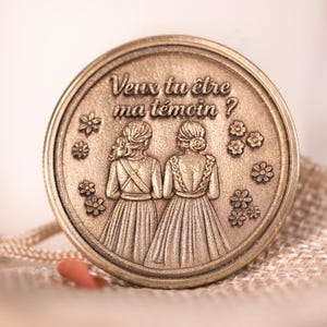 Peut inclure: Une pièce ronde de couleur bronze avec le texte "Veux tu être ma témoin ?" et une image en relief de deux femmes en robes, entourées de petites fleurs. Il s'agit probablement d'un cadeau de mariage.