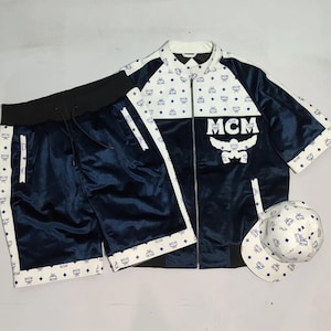 May include: A navy blue tracksuit set featuring a zip-up jacket, shorts, and a baseball cap. The jacket and cap have a white pattern with the letters "MCM". The shorts have a black waistband and white trim.