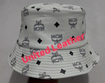 May include: White bucket hat featuring a repeating pattern of black MCM logos and diamond shapes. The hat is made of a smooth material and has a classic, casual style. The hat is a fashion accessory.