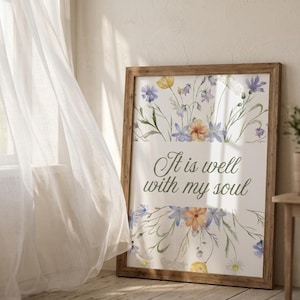 May include: A framed print with the text "It is well with my soul". The artwork features watercolour flowers in yellow, blue, and orange. The frame is made of light brown wood, adding a rustic touch. The print is displayed indoors.