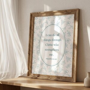 May include: A framed print with a light brown wooden frame. The print has a floral pattern in light blue on a cream background. An oval in the centre contains the text "I can do all things through Christ who strengthens me." from Philippians 4:13.