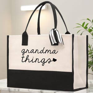 Puede incluir: Un bolso tote grande con asas negras y una base negra. La bolsa es principalmente blanquecina con las palabras "grandma things" impresas en escritura negra, junto con un pequeño corazón negro. Una cinta a rayas blancas y negras está atada a una de las asas.