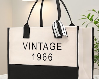 Op de afbeelding: Een crèmekleurige draagtas met zwarte details. De onderkant en handvatten zijn zwart, terwijl de hoofdgedeelte de tekst "VINTAGE 1966" in zwart weergeeft. Een zwart-wit gestreept lint is aan een van de handvatten geknoopt.