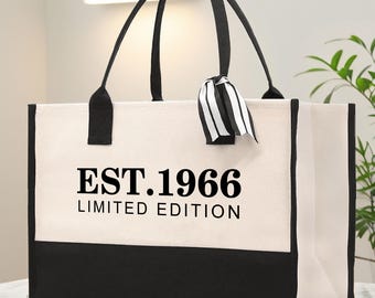 Op de afbeelding: Een rechthoekige canvas tas met een crèmekleurig bovengedeelte en een effen zwarte onderkant. De voorzijde toont de zwarte tekst EST.1966 LIMITED EDITION. De tas heeft twee zwarte hengsels en een decoratief zwart-wit gestreept lint.
