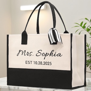 May include: A large tote bag with black handles and a black base. The bag is off-white with the text "Mrs. Sophia EST 10.28.2025" printed on the front. A black and white striped ribbon is tied to one of the handles.