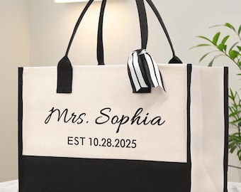 May include: A large tote bag with black handles and a black bottom panel. The bag is off-white with the text "Mrs. Sophia EST 10.28.2025" printed on the front. A black and white striped ribbon is tied to one of the handles.