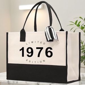 Könnte beinhalten: Eine cremefarbene Canvas-Tragetasche mit schwarzen Henkeln und einem schwarzen Boden. Die Tasche trägt den schwarzen Text "LIMITED 1976 EDITION". Ein schwarz-weiß gestreiftes Band ist an einem der Griffe befestigt.