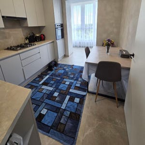 May include: A rectangular area rug featuring a patchwork design of various shades of blue and brown denim fabric squares. The rug is positioned on a light-colored tile floor in a kitchen and dining area, with a small black dog resting on its edge.