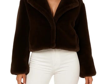May include: A dark brown faux fur jacket with a wide collar and long sleeves. The jacket is cropped and has a button closure. The model is wearing white pants.