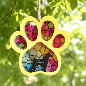 May include: A yellow paw-shaped ornament with a stained-glass design. The ornament features a black silhouette of a dog with the name "Kaylan" and is suspended by a twine string. The stained glass has a colorful mosaic pattern.