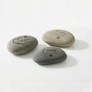 May include: Three engraved memorial stones. One stone has a heart and the name "Melina" with dates. Another stone has the name "Kitty" and dates. The third stone has the name "Cindy" and dates. Each stone has a paw print.