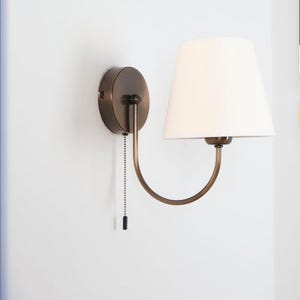 Può includere: Una lampada da parete con base circolare in bronzo e un braccio curvo che sostiene un paralume in tessuto bianco di forma conica. Una sottile catenella nera pende dalla base e funge da interruttore manuale per la luce.