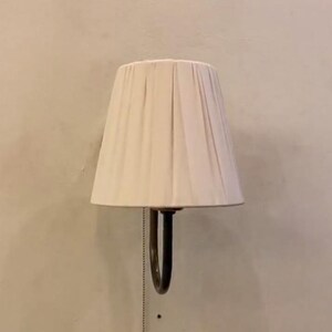 May include: A wall-mounted lamp featuring a cream-colored, pleated fabric shade with a tapered silhouette. The fixture includes a dark metal curved arm and a vertical pull chain with a small circular ring at the bottom for manual operation.