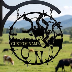 May include: Black metal sign featuring two goats within a barbed wire circle. The sign includes the text "CUSTOM NAME" and initials "C N". The sign is hanging on a wooden post with a green pasture and mountains in the background.