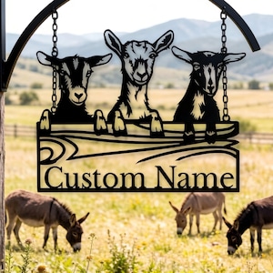 May include: Black metal sign depicting three goat silhouettes above a wooden sign with the text "Custom Name". The sign is suspended from a curved bracket with chains. Three donkeys graze in a grassy field below.