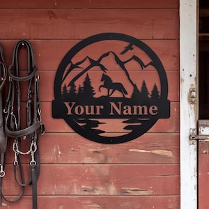 May include: A circular black metal wall sign featuring a cutout silhouette of a horse, pine trees, mountains, and a bird in flight. A horizontal bar across the center contains the text Your Name in a serif font, centered within the circular frame.