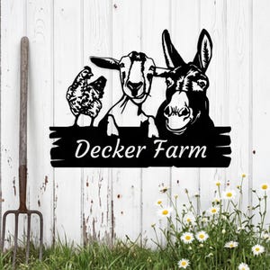 Puede incluir: Letrero de metal negro con una gallina, una cabra y un burro. El letrero dice "Decker Farm" en letras blancas. Está montado en una valla de madera blanca, con una horca y margaritas en primer plano.