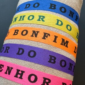 Può includere: Primo piano di braccialetti in tessuto colorati. Ogni fascia è di un colore diverso: blu, giallo, arancione, viola e rosa. Le fasce hanno un testo nero che recita "DO BON", "NHOR DO", "BONFIM", "OR DO BON" e "ENHOR DO".