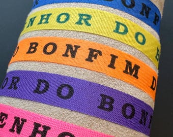 Op de afbeelding: Een close-up van kleurrijke stoffen polsbandjes. Elke band heeft een andere kleur: blauw, geel, oranje, paars en roze. De bandjes hebben zwarte tekst met de tekst "DO BON", "NHOR DO", "BONFIM", "OR DO BON" en "ENHOR DO".