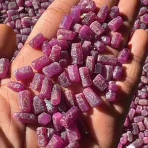 Könnte beinhalten: Nahaufnahme einer Hand, die rohe, rosa Rubine hält. Die unregelmäßig geformten Edelsteine haben eine intensive rosa Farbe. Der Hintergrund ist mit weiteren Steinen gefüllt, was einen strukturierten Effekt erzeugt.
