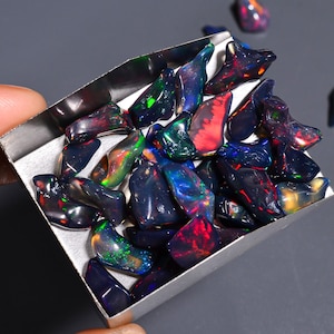 May include: A selection of polished black opal gemstones, displaying a spectrum of colours such as red, green, and blue. The opals are presented in a small, silver tray, highlighting their unique forms and iridescent characteristics. The backdrop is a neutral grey.