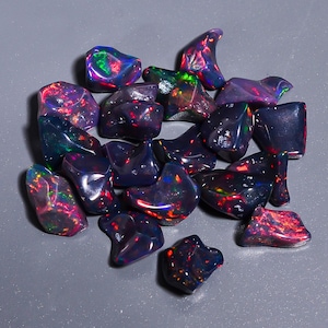May include: A collection of polished black opal gemstones with vibrant flashes of color, including red, green, and blue. The opals are irregularly shaped and sit on a gray surface. These gemstones are ideal for jewelry making.