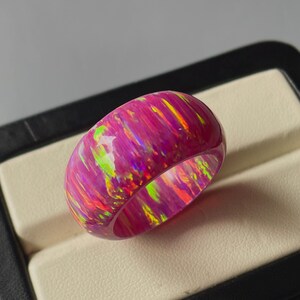 Può includere: Un anello a fascia larga con finitura lucida. L'anello presenta un mix di colori rosa, verde e giallo, creando un effetto iridescente unico. L'anello è presentato su una superficie dai toni neutri.
