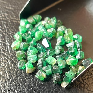 May include: A collection of raw, uncut emerald gemstones. The emeralds are a vibrant green color, with varying shapes and sizes. They are displayed on a dark, textured surface, possibly for sorting or presentation.