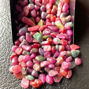 May include: A collection of polished gemstones in shades of red, pink, and green, spilling from a metal container. The stones vary in size and shape, with a smooth, rounded texture. The gemstones are scattered on a dark surface.