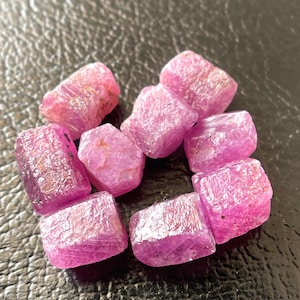 May include: A cluster of raw ruby gemstones, displaying a vivid pink-red hue. The stones have a rough, textured appearance, suggesting they are uncut. The gemstones are irregularly shaped, showcasing their natural crystal form.