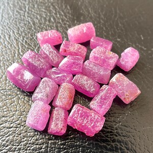 May include: A collection of raw, uncut ruby gemstones. The stones are a vibrant pinkish-red color, with a rough, textured surface. The gemstones are scattered across a dark surface.