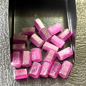 Może przedstawiać: Kolekcja fasetowanych, różowych kamieni szlachetnych o prostokątnym kształcie. Kamienie szlachetne są prezentowane w czarnej tacy, ukazując ich żywy kolor i krystaliczną strukturę. Odcienie wahają się od głębokiej magenty do jasnoróżowego.