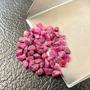 Op de afbeelding: Een verzameling ruwe, ongeslepen robijn edelstenen in verschillende tinten roze en rood. De edelstenen liggen op een kleine, zilveren metalen bak, tegen een getextureerde, donkere ondergrond. De ruwe robijnen hebben een natuurlijke, onregelmatige vorm.