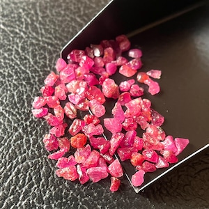 Peut inclure: Un ensemble de pierres précieuses rubis brutes et non taillées. Les petites pierres de forme irrégulière sont d'un rouge-rose vif. Elles sont présentées sur une surface noire, partiellement contenues dans une petite pelle noire.
