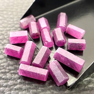 May include: A collection of small, elongated, hexagonal-shaped pink gemstones with a translucent, crystalline texture. The stones are scattered and clustered together, resting partially inside a black metal scoop with a smooth, angled edge.