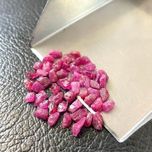 Op de afbeelding: Een verzameling kleine, ruw geslepen robijn edelstenen in verschillende tinten roze en rood. De edelstenen liggen op een donkere ondergrond naast een kleine roestvrijstalen bak. Een enkel wit object bevindt zich tussen de edelstenen.