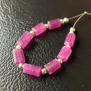 Może przedstawiać: Sznur żywych, różowych koralików z kamieni szlachetnych rubinu. Każdy prostokątny koralik jest oddzielony małym, srebrnym koralikiem. Kamienie szlachetne mają lekko przezroczystą jakość, ujawniając wewnętrzne tekstury. Sznurek jest widoczny na końcach.