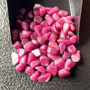 Puede incluir: Una colección de piedras preciosas de rubí pulidas y pulidas en un pequeño recipiente negro. Las piedras preciosas son de un color rojo rosado vibrante, con formas lisas y redondeadas. El recipiente está inclinado, derramando las piedras preciosas sobre una superficie negra texturizada.