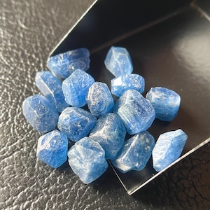 Puede incluir: Un conjunto de pequeñas gemas azules facetadas. Las piedras son de un color azul claro vibrante y parecen ser en bruto. Se encuentran dentro de una bandeja oscura en ángulo, lo que sugiere que se están exhibiendo o clasificando.