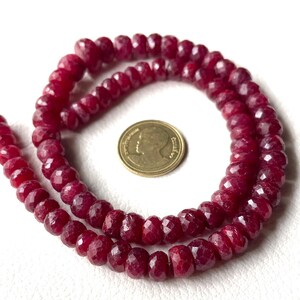 May include: A necklace of faceted, deep red ruby beads. The beads are arranged in a circular pattern, with a gold coin placed in the centre for size comparison. The beads have a polished, reflective surface, and are strung together.