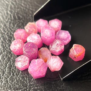 May include: A collection of raw, uncut ruby gemstones. The rubies display a vibrant pink hue, with some stones exhibiting a deeper, more saturated color. The rough texture and irregular shapes of the stones are visible.