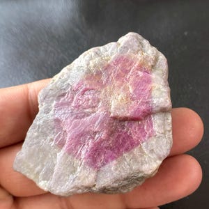 Puede incluir: Una gema en bruto con una mezcla de colores. La piedra tiene una base de gris claro y blanco, con un área prominente de tonos rosa y magenta vibrantes. La textura parece áspera y natural.