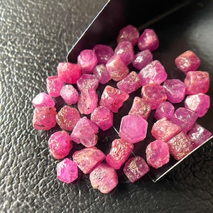Könnte beinhalten: Eine Sammlung roher, ungeschliffener Rubin-Edelsteine. Die Rubine variieren in Größe und Form und zeigen eine Reihe von rosa und roten Farbtönen. Einige Steine haben eine durchscheinende Qualität, andere erscheinen undurchsichtiger. Die Edelsteine liegen auf einer strukturierten schwarzen Oberfläche.