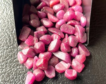 Peut inclure: Une collection de pierres précieuses rubis polies et roulées, de différentes nuances de rose et de rouge. Les pierres sont lisses et de forme irrégulière, présentées dans un petit récipient sombre sur une surface noire texturée.