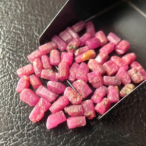 Peut inclure: Collection de pierres précieuses de rubis bruts et non taillés. Les pierres précieuses sont d'un rouge-rosé vif et ont une texture brute et naturelle. Elles sont contenues dans un petit récipient sombre, avec quelques-unes qui en débordent.
