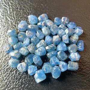 May include: A collection of small, faceted blue gemstones. The stones have a translucent quality with some showing internal inclusions. The gems are scattered on a dark, textured surface, creating a contrast that highlights their color and form.
