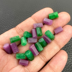 Peut inclure: Une collection de petites perles de pierres précieuses brutes et cylindriques dans des tons vifs de vert émeraude et de rose magenta profond. Ces pierres non polies présentent une section hexagonale et une texture mate naturelle.