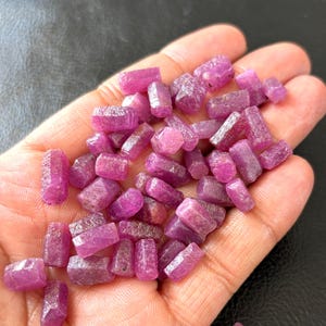Könnte beinhalten: Nahaufnahme einer Handvoll roher, ungeschliffener Rubinkristalle. Die Kristalle sind tiefrosa-rot und haben eine raue, natürliche Textur. Die Edelsteine sind klein, etwa 0,6 bis 1,9 cm lang, und werden in einer offenen Hand gehalten.